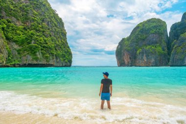 Tayland, Krabi 'deki Phi Phi adalarında turkuaz su okyanusuyla tropik kumlu Maya Koyu' nun deniz kıyısında duran bir gezgin. Boş plajda kayalıklı erkek turistin arka görüntüsü
