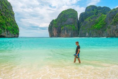 Tayland, Krabi 'deki Phi Phi adalarında turkuaz su okyanusuyla tropik kumlu Maya Koyu' nun deniz kıyısında yürüyen bir gezgin. Dağları ve yeşil bitkileri olan boş plajda erkek turist