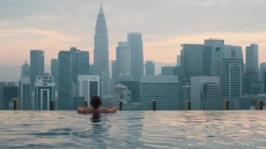 Kuala Lumpur, Malezya - 7 Haziran 2024: Erkek turist güneş doğarken sonsuz çatı üstü yüzme havuzunda dinleniyor, Kuala Lumpur. Şehir merkezi finans ve iş merkezi ile şehir manzarasının keyfini çıkaran erkek gezgin