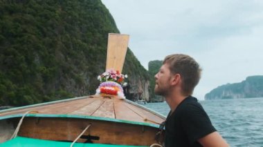 Tayland 'da Phi Phi adalarında turkuaz suyu ve yeşil kayalıkları olan geleneksel Tayland ahşap uzun kuyruklu teknesinin pruvasında oturan erkek turist. Maya Körfezi sahiline yelken açıyorum. Asya 'da seyahat ve turizm.