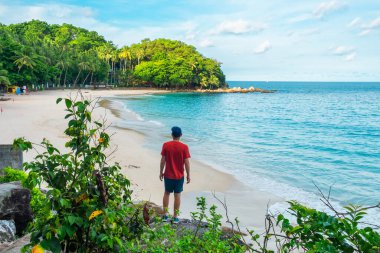 Bir tepede duran turistin arka görüntüsü, tropikal kumlu sahile bakan temiz mavi su okyanusu ve yemyeşil arazisi olan. Tayland, Phuket Adası 'nda Özgürlük Sahili' nin üzerinde duran erkek yolcu