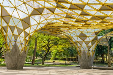 Perdana Botanik Bahçesi, Kuala Lumpur, Malezya, modern mimari yapısı ile sarı geometrik çatısı ve geniş açık alanı ile çevreleyen yeşil ve parlak mavi gökyüzü altında ağaçlar.