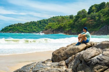 Erkek gezgin, tropik kumlu sahildeki kayalıklarda turkuaz su okyanusuyla oturur. Tayland 'ın Phuket Adası' ndaki Özgürlük Sahili 'nde yalnız başına dinlenen erkek turist. Kış ve yaz tatilleri
