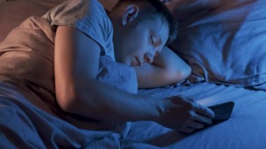 Beyaz bir adam cep telefonuyla evde uyuyakalmış. Yorgun ve bitkin bir erkek yatak odasında akıllı telefonuyla uyuyor. Akıllı telefon, internet, sosyal medya, alet bağımlılığı kavramı.