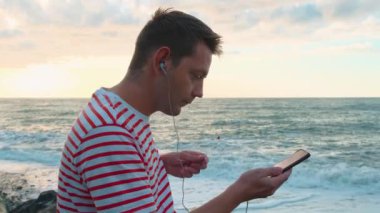 Deniz kıyısında kulaklık takan ve müzik dinleyen akıllı telefonlu neşeli beyaz bir genç. Yakışıklı erkek gün batımında plajda kulaklıklı cep telefonu kullanıyor..