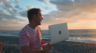 Gün batımında Rocky Beach 'te dizüstü bilgisayarı olan genç bir beyaz adam video görüşmesiyle çevrimiçi sohbet ediyor ve dizüstü bilgisayar kamerasıyla Altın Deniz Günbatımını gösteriyor. Dijital göçebe yaşam tarzı. İş-yaşam dengesi.