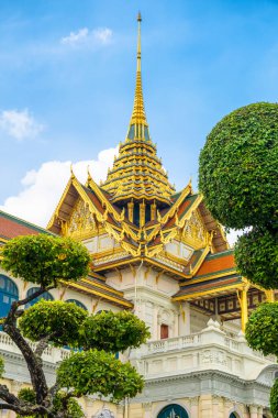 Bangkok, Tayland 'daki Grand Palace' da altın çatılı geleneksel Tayland mimarisi, süslü detaylar ve parlak mavi gökyüzüne karşı manikürlü yeşil ağaçlar var. Dikey oryantasyon. Seyahat ve