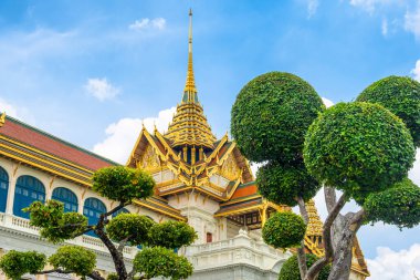Bangkok, Tayland 'daki Grand Palace' da altın çatılı geleneksel Tayland mimarisi, süslü detaylar ve parlak mavi gökyüzüne karşı manikürlü yeşil ağaçlar var. Asya 'da seyahat ve turistik dönüm noktası.
