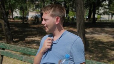 Sıcak hava dalgasından muzdarip bir adam. Beyaz erkek, dışarıda otururken serinlemek ve susuzluktan kurtulmak için su içiyor. Yorgun ve hararetli bir adam şehrin dışındaki bankta oturuyor..