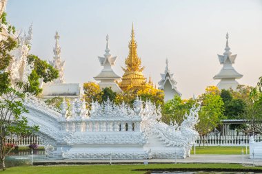 Gün batımında ağaçlar ve yeşilliklerle çevrili altın ve beyaz Budist tapınak yapıları, Chiang Rai, Tayland. Wat Rong Khun kompleksi. Asya 'da seyahat ve turistik dönüm noktası.