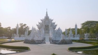 Karmaşık oymaları olan beyaz tapınak, su birikintisi üzerinde köprü ve gün batımında yeşil bahçe, Chiang Rai, Tayland. Kuzey Tayland 'daki Budist Wat Rong Khun tapınağına giriş. Seyahat simgesi.