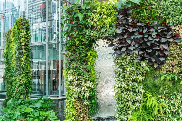Kentsel mimari ortamda modern cam şelaleyi çevreleyen çeşitli bitkilerle yemyeşil bir dikey bahçe. Bangkok 'taki alışveriş merkezine entegre edilmiş yeşil tesisleri olan duvarlar.