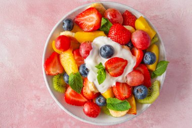 Bir kase renkli meyve salatası ve pembe arka planda kremalı yoğurt. Üst Manzara. Vitaminli sağlıklı vejetaryen yemeği. Karışık çilek, üzüm, muz, kivi, yabanmersini, şeftali, diyet öğle yemeği için nane..