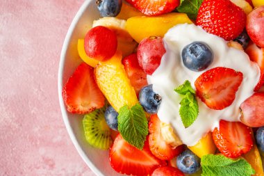 Yaz mevsiminin renkli meyve salatasını pembe arka planda kremalı yoğurtla kapat. Üst Manzara. Vitaminli sağlıklı vejetaryen yemeği. Karışık çilek, üzüm, muz, kivi, yabanmersini, şeftali, nane