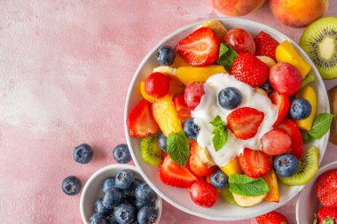 Yazın pembe arka planda kremalı yoğurdun içinde renkli meyve salatası. Kopyalama alanı olan üst görünüm. Kahvaltıda sağlıklı vitamin yiyeceği. Karışık çilek, üzüm, muz, kivi, yabanmersini, şeftali, nane