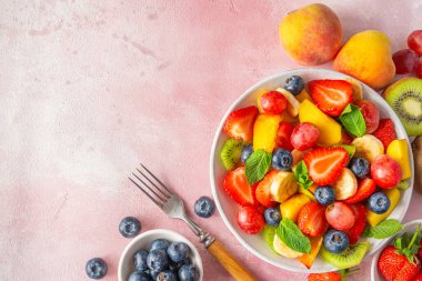 Pembe arka planda çatallı bir kasede renkli meyve salatası. Kopyalama alanı olan üst görünüm. Kahvaltıda sağlıklı vitamin yiyeceği. Karışık çilek, üzüm, muz, kivi, yabanmersini, şeftali, nane