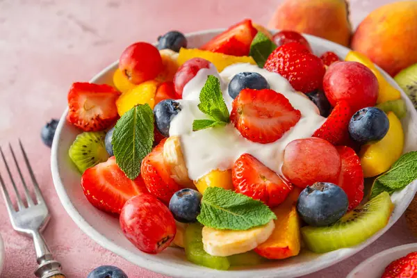 Yaz mevsiminin renkli meyve salatasını, pembe arka planda Yunan yoğurdu kasesiyle kapat. Vitaminli sağlıklı vejetaryen yemeği. Karışık çilek, üzüm, muz, kivi, yabanmersini, şeftali, diyet için nane.