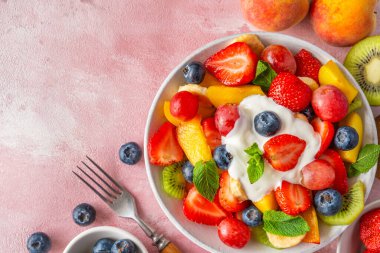 Yazın pembe arka planda çatallı kremalı yoğurt ile renkli meyve salatası. Kopyalama alanı olan üst görünüm. Kahvaltıda sağlıklı vitamin yiyeceği. Karışık çilek, üzüm, muz, kivi.