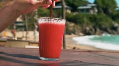 Erkek el, Tayland 'ın Samui adasındaki sahil kafesinde, tahta bir masa üzerinde cam içinde ferahlatıcı kırmızı karpuz smoothie karıştırıyor. Coral Cove 'daki yaz restoranında soğuk tropik meyve suyu.