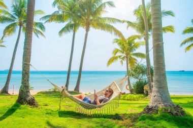 Deniz kenarındaki palmiye ağaçlarının altında uzanan adam, tropik plaj tatilinin tadını çıkarıyor güneşli bir günde dinleniyor ve akıllı telefon kullanıyor, okyanus kenarında huzurlu bir tatil anı, Phu Quoc Adası, Vietnam. Yaz