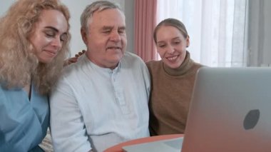 Yaşlı bir baba, iki yetişkin kızıyla birlikte oturmuş dizüstü bilgisayardan ona sarılıyor. Aşk, ilgi, destek ve aile bağı, modern aile teknolojisi kullanımı, duygusal bağlantılar.