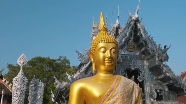 Tayland 'ın Chiang Mai şehrinde, açık mavi gökyüzü altında, karmaşık ejderha oymaları ve detaylı metal işleriyle süslü Gümüş Tapınak' ın önündeki altın Buda heykeli. Wat Sri Suphan Budist Tapınağı. Asya
