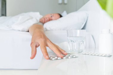 Hasta adam yatağında uzanıp bir bardak suyla komodinin üzerinde ilaç almaya çalışıyor. Sağlık, grip tedavisi, sağlıklı alışkanlıklar, stres atma, hastalık, kendi kendine tedavi, virüs ve enfeksiyon konsepti.