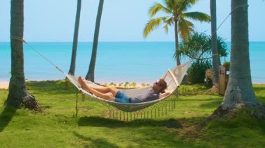 Turkuvaz deniz ve yeşil çimen ile tropik sahildeki palmiye ağaçlarının arasında dinlenen bir adam, rahatlama, yaz tatili ve huzurlu bir kaçış için cennet, Phu Quoc Adası, Vietnam