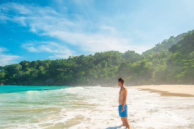 Tropikal sahilde köpüklü dalgalarda tek başına duran bir adam. Turkuaz dalgalar ve Tayland 'ın Phuket adasında yemyeşil bir orman. Özgürlük plajında yalnız erkek turist. Popüler yaz tatili merkezi