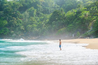 Adam, Tayland 'ın Phuket Adası' ndaki turkuaz dalgalar ve yemyeşil ormanlarla tropik sahillerdeki sığ dalgalarda tek başına sörf yapıyor. Özgürlük plajında yalnız erkek turist. Popüler yaz tatili
