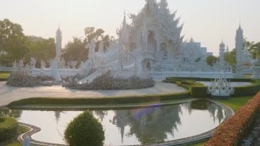 White temple with intricate carvings, reflection in water pond and green garden at sunset, Chiang Rai, Thailand. Entrance to Buddhist Wat Rong Khun temple in Northern Thailand. Travel landmark.