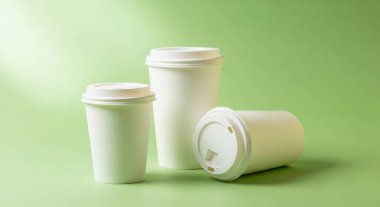 Three disposable white paper coffee cups with lids are arranged on a green background. The cups are different sizes and one is lying on its side. Studio shot. 150 chars