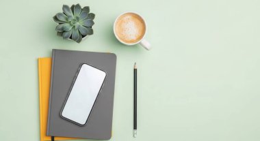 Overhead shot of a workspace with a smartphone, cup of coffee, succulent, notebooks and pencil on a light green background. Ideal for blog or website.