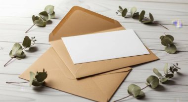 An overhead shot showcases a blank card nestled in a brown envelope, adorned with eucalyptus leaves on a white wooden table. Perfect for invitations and greetings.