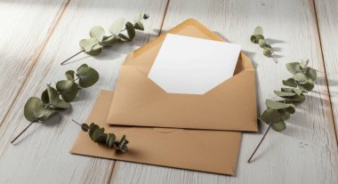 A blank card with a brown envelope and eucalyptus leaves is placed on a white wooden table. Perfect for adding your message, invitation, or greeting. Flat lay composition.