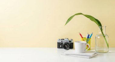 Antika bir kamera, bir fincan kahve ve bir bitki beyaz bir masada sarı arka planda sıralanır. Bu görüntü bir nostalji ve yaratıcılık hissi uyandırıyor..