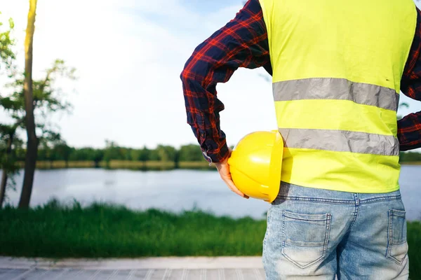 Construction worker wearing yellow vest staying in front of the lake. Worker holding yellow helmet.