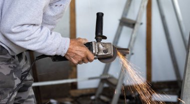 Yaşlı bir işçi metal öğütür. Yaşlı bir adam elinde bir öğütücü tutuyor ve kıvılcımlar uçuşurken metal bir boruyu öğütüyor. Öğütücüyü tutan el odaklanmış durumda.