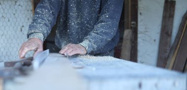 Yaşlı bir marangoz dairesel bir testereyle odun kesiyor. İşçinin elleri odaklanmış. Bir eliyle tahta bir gövde tutuyor, diğer eliyle de ihtiyacı olan aleti alıyor. Etrafta talaş var.