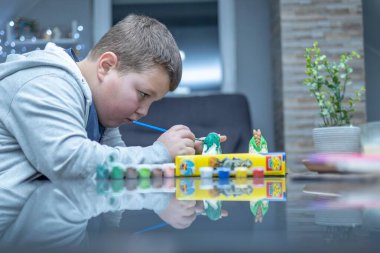 a boy is coloring a dinosaur figure. he is very focused and serious. he holds a paint brush and carefully applies paint to the figure