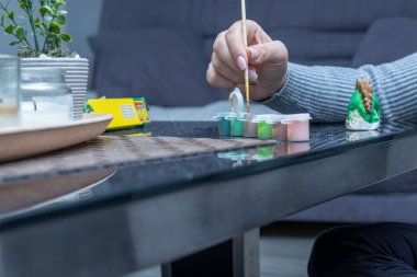 a woman's hand holding a paintbrush and dipping it in paint. a woman paints a ceramic dinosaur figure. on the table are paints, a dinosaur figure and ceramic particles that fell off the figure