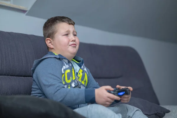 the boy is sitting on the couch, holding a joystick and playing a video game.he is sad because he cannot complete the mission.the boy almost cried.honest emotion of a boy playing a video game