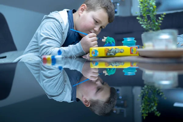a boy paints a ceramic dinosaur figure. he holds a paintbrush and carefully brings it to the figure. he looks at the figure and the brush. interesting photo of the boy's reflection on the glass table
