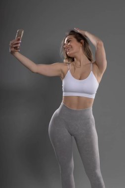 Genç çekici fitness kızı gri arka planlı stüdyoda poz veriyor ve selfie çekiyor.