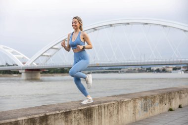 Spor giyimli genç bir kadın iskelede koşuyor ve gülümsüyor. Sağlıklı yaşam tarzı, spor yaşam tarzı. Arka planda bir köprü var.