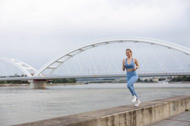 Spor giyimli genç bir kadın iskelede koşuyor ve gülümsüyor. Sağlıklı yaşam tarzı, spor yaşam tarzı. Arka planda bir köprü var.
