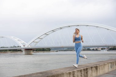 Spor giyimli genç bir kadın iskelede koşuyor ve gülümsüyor. Sağlıklı yaşam tarzı, spor yaşam tarzı. Arka planda bir köprü var.