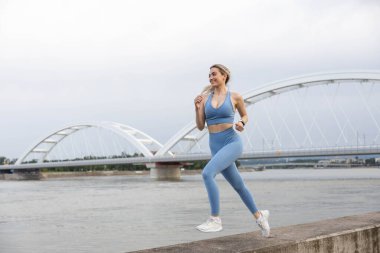 Spor giyimli genç bir kadın iskelede koşuyor ve gülümsüyor. Sağlıklı yaşam tarzı, spor yaşam tarzı. Arka planda bir köprü var.