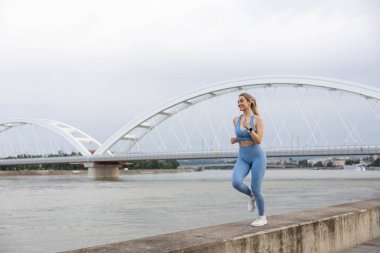 Spor giyimli genç bir kadın iskelede koşuyor ve gülümsüyor. Sağlıklı yaşam tarzı, spor yaşam tarzı. Arka planda bir köprü var.