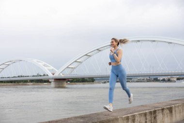 Spor giyimli genç bir kadın iskelede koşuyor ve gülümsüyor. Sağlıklı yaşam tarzı, spor yaşam tarzı. Arka planda bir köprü var.